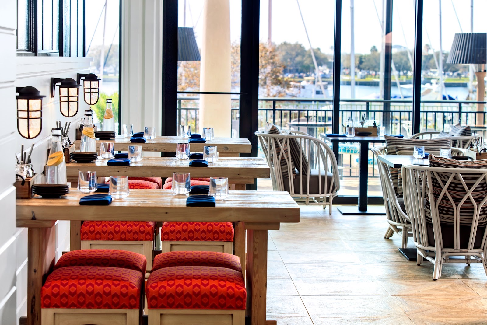 The dining room: reclaimed-wood high tables with red woven stools and white rattan chairs under black lantern sconces, the marina through tall windows