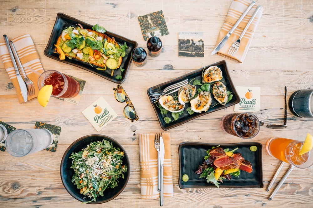 An overhead spread of plates on a pale wood table: a seafood toast, charred oysters, a chopped salad and seared tuna, with Paul’s Landing coasters