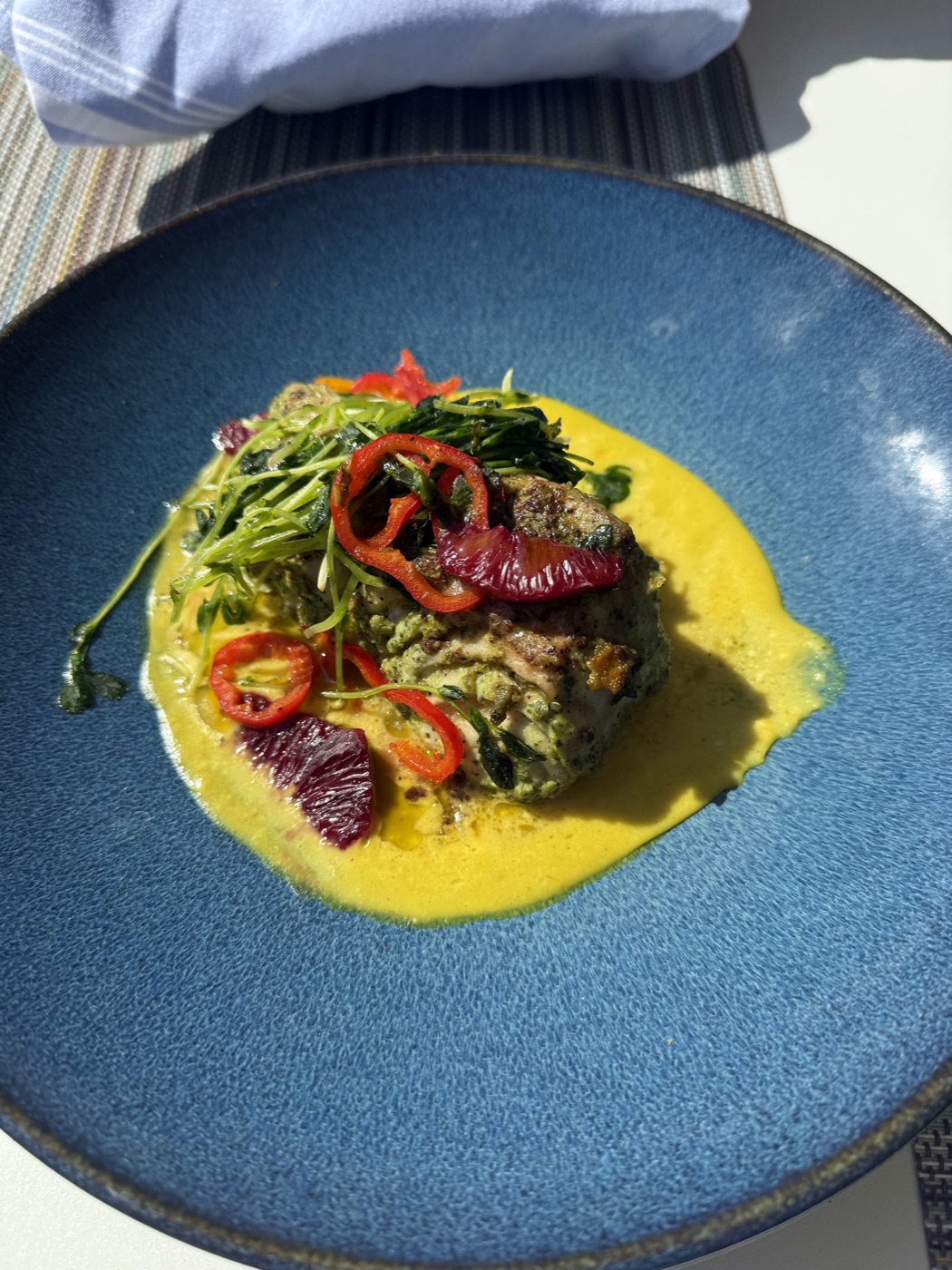 A herb-crusted grouper fillet over yellow curry sauce on a blue plate, topped with red chilies, blood orange and pea shoots