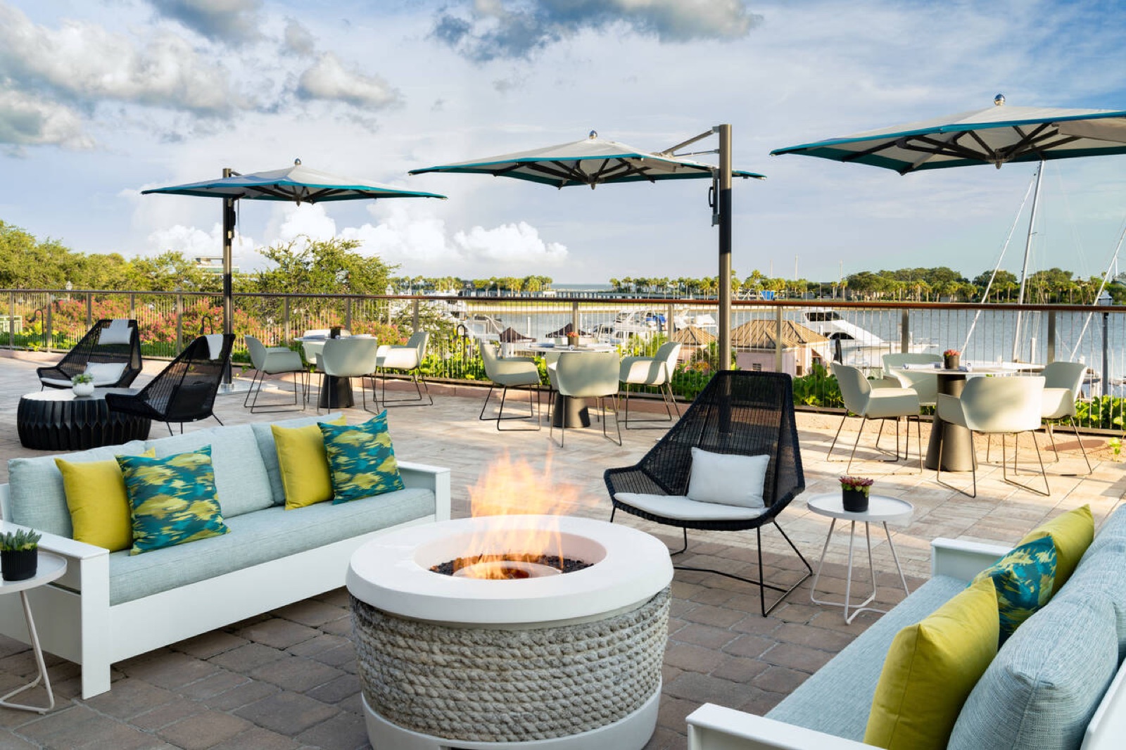 The patio at Paul’s Landing: cushioned sofas with yellow pillows around a lit fire pit, teal-and-white umbrellas, and the marina beyond